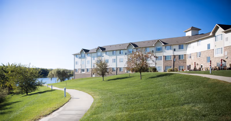 A large senior living facility building with multiple windows and a brick and siding exterior, situated on a grassy hill with a paved walkway leading towards a lake in the background under a clear blue sky.