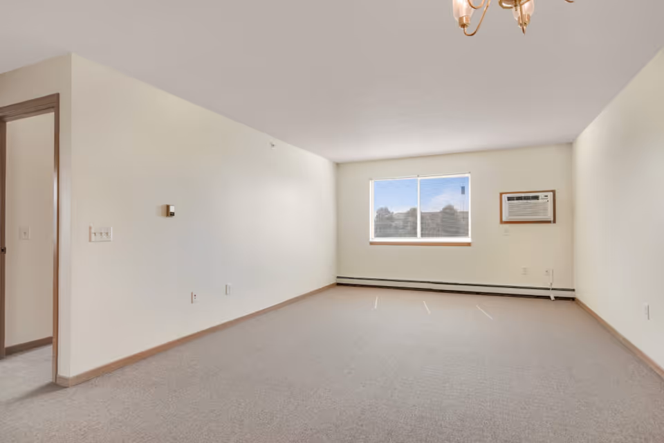 Empty room with beige carpet, cream-colored walls, a window with blinds showing trees outside, a wall-mounted air conditioning unit, and a doorway leading to another room.