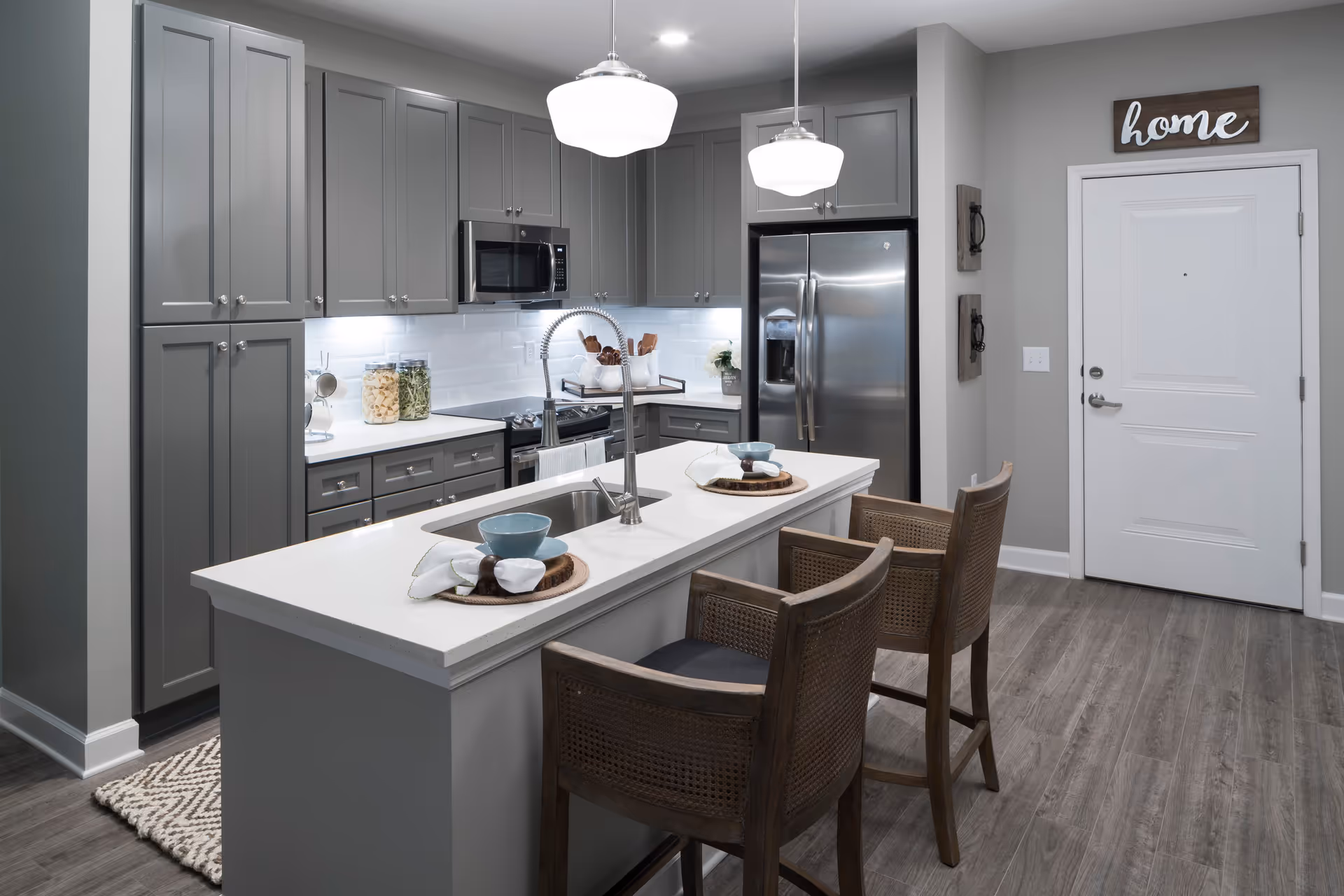 Modern kitchen with gray cabinets, stainless steel refrigerator and microwave, white countertops, a kitchen island with a sink, two wooden chairs, and two pendant lights hanging above the island. A white door with a wooden sign that says 'home' is visible in the background.