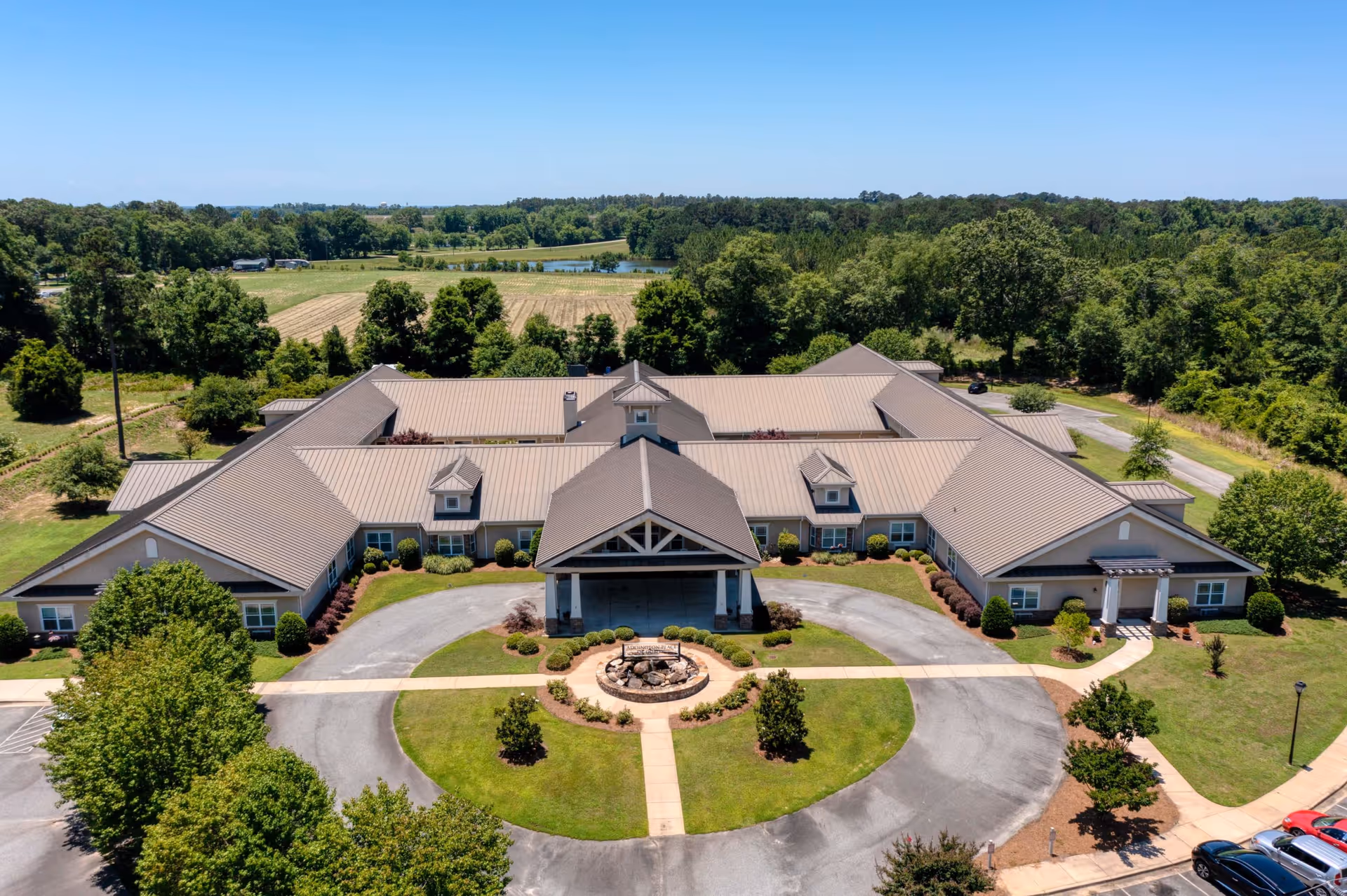 Aerial view of Addington Place of Dublin, a single-story senior living facility with a large circular driveway and landscaped greenery. The building is surrounded by trees and open fields under a clear blue sky.