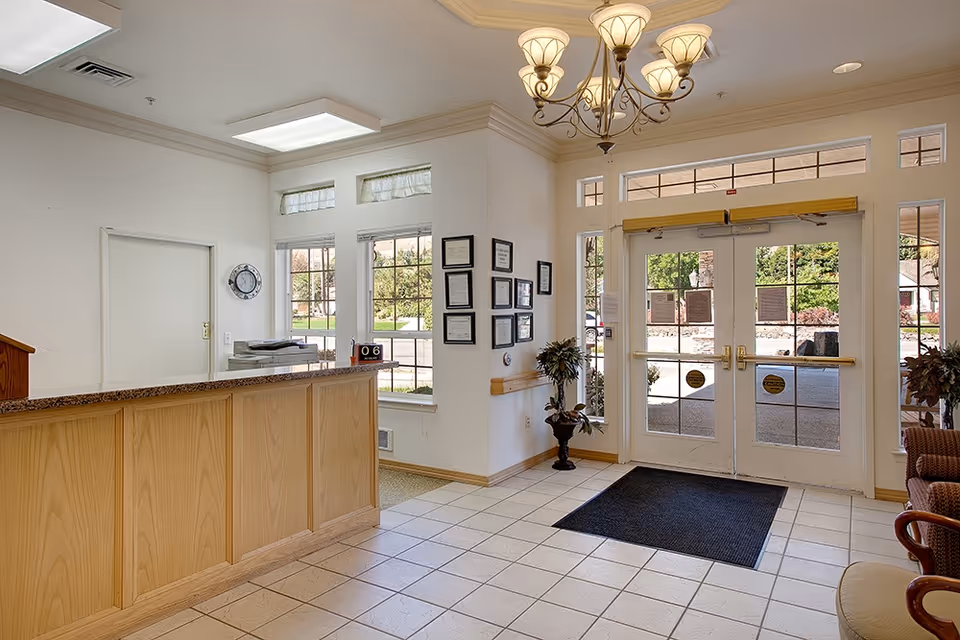 Reception area of a senior living facility with a wooden front desk, a clock on the wall, framed certificates, potted plants, a chandelier, and glass double doors leading outside.