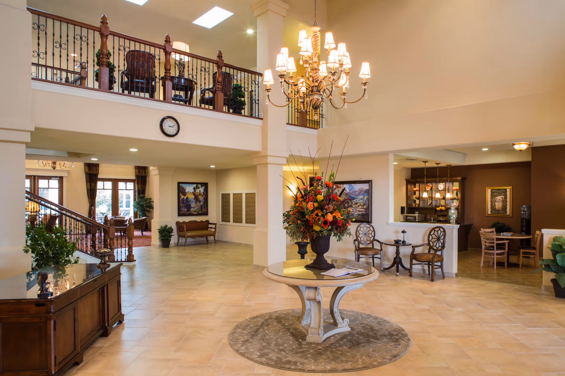 Spacious and well-lit senior living facility lobby with a large floral arrangement on a round table in the center. The area features a chandelier, a staircase with wooden railings, seating areas with chairs and tables, and decorative paintings on the walls. There is a balcony overlooking the lobby and a clock mounted on the wall.