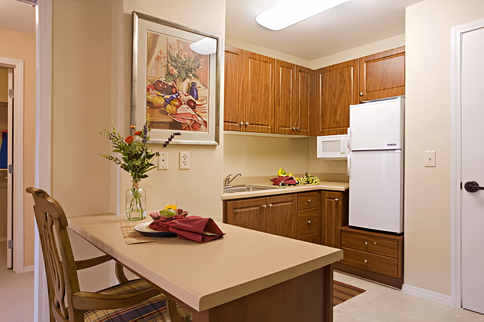 Small kitchen with wooden cabinets, a white refrigerator, a countertop island with a place setting and vase of flowers.