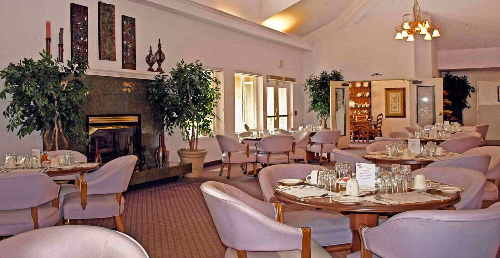 A dining room with round tables set with plates, cups, and glasses. The room features lavender upholstered chairs, a fireplace with decorative items above it, potted plants, and a chandelier hanging from the ceiling. There are windows and double doors letting in natural light.