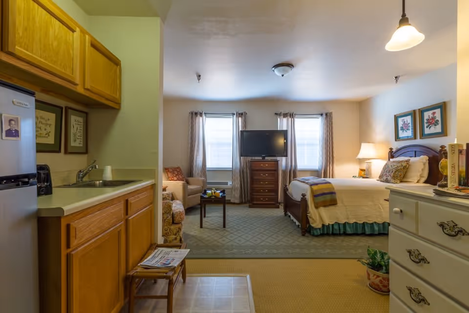 A cozy assisted living studio apartment featuring a small kitchenette with wooden cabinets and a stainless steel refrigerator on the left. The room opens into a living and sleeping area with a bed, a wooden dresser with a TV on top, two windows with curtains, an armchair, a small coffee table, and framed floral artwork on the walls. A lamp on a bedside table provides warm lighting.