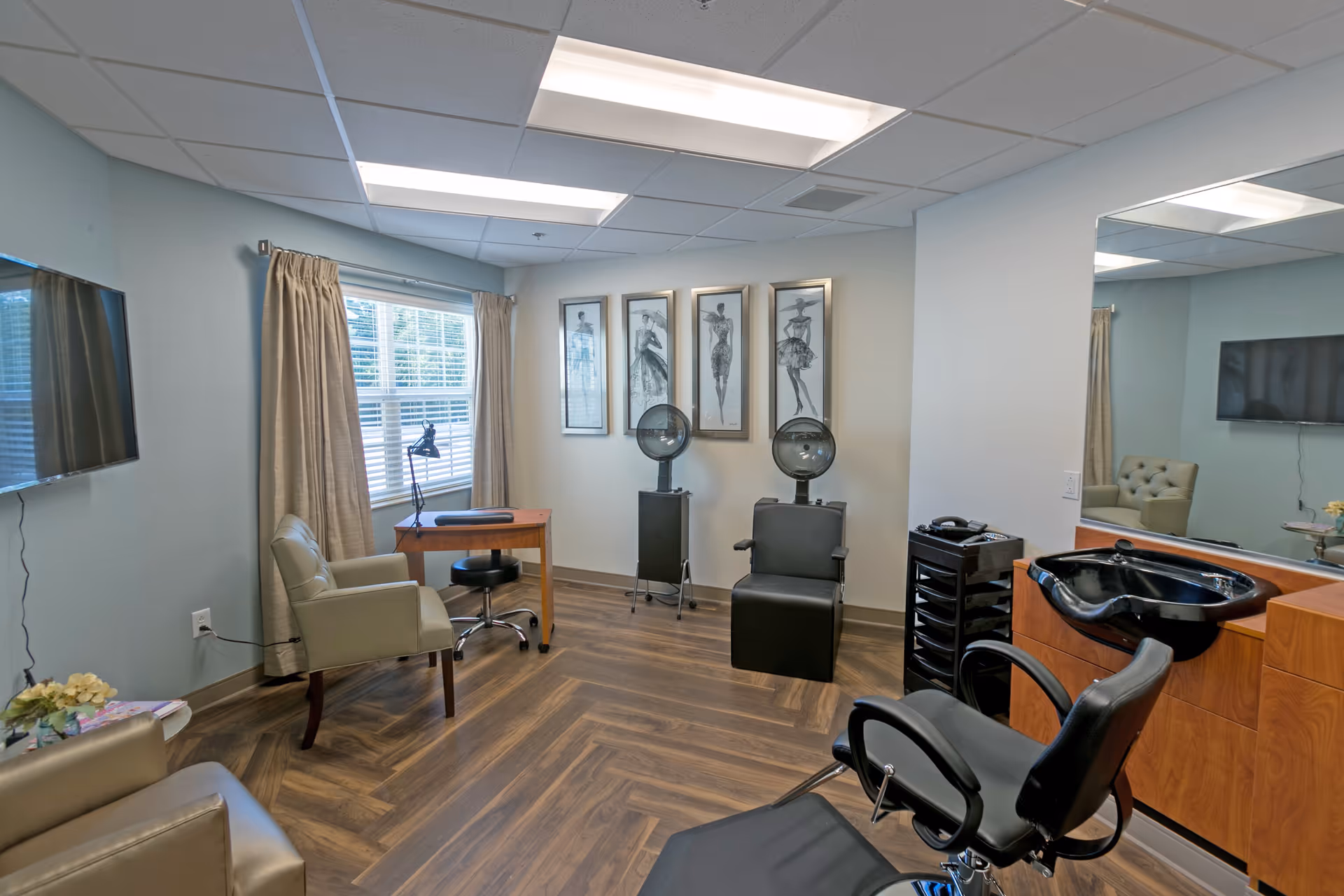 Interior of a salon room with a black salon chair and hair washing station, two hair dryers on stands, a small wooden desk with a lamp, two beige armchairs, a wall-mounted TV, and four framed fashion sketches on the wall.