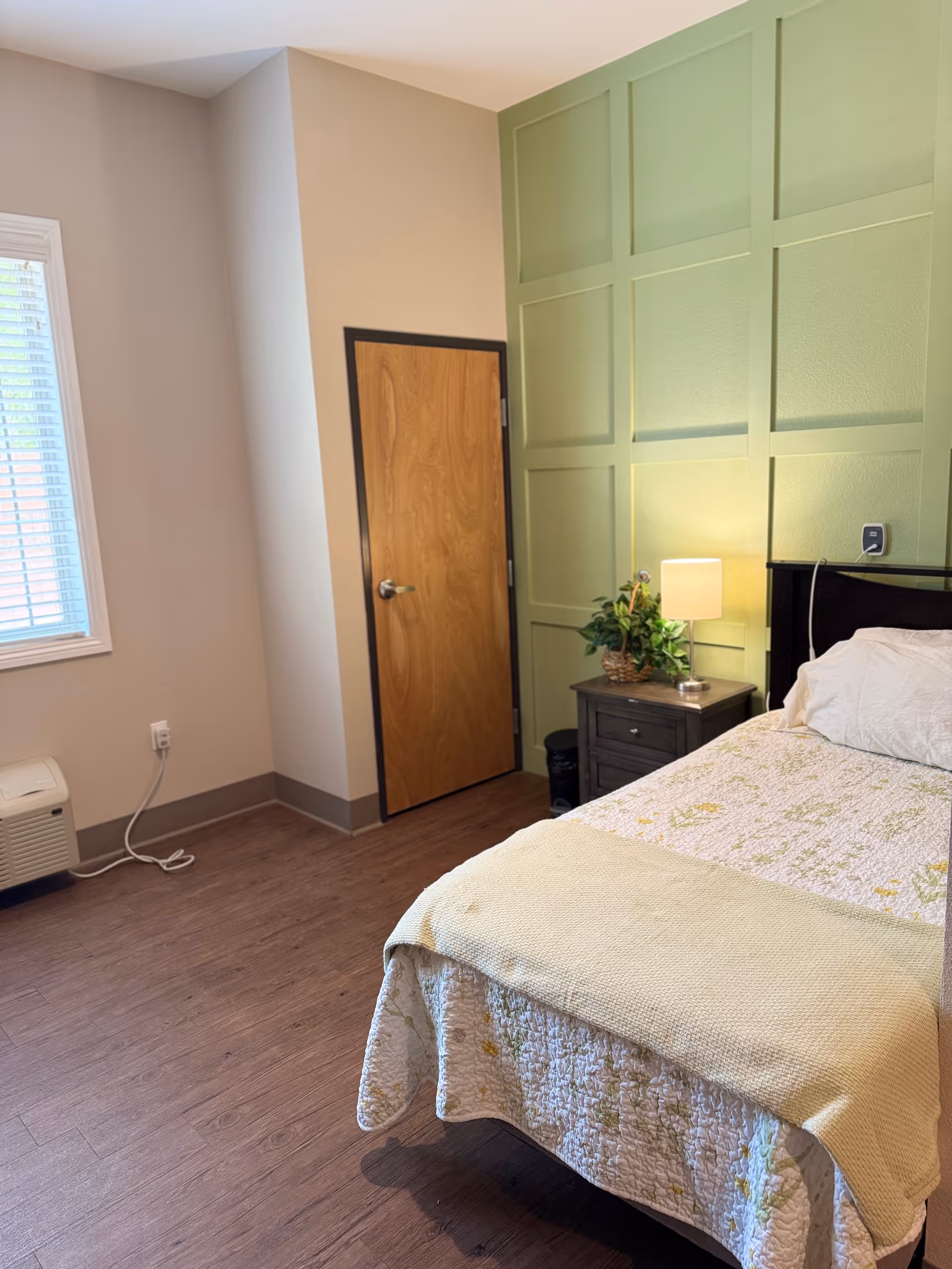 A cozy bedroom with a single bed covered in a floral quilt and a light yellow blanket. Next to the bed is a dark wooden nightstand with a small lamp and a potted plant. The wall behind the bed is painted green with a decorative panel design. There is a wooden door with a silver handle and a window with white blinds letting in natural light. The floor is a dark wood laminate.