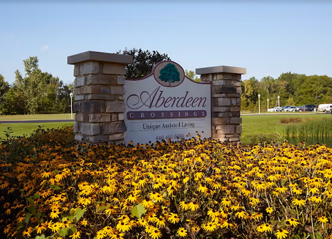 Stone sign for Aberdeen Crossings Unique Assisted Living surrounded by a large bed of yellow flowers with green trees and a clear blue sky in the background.