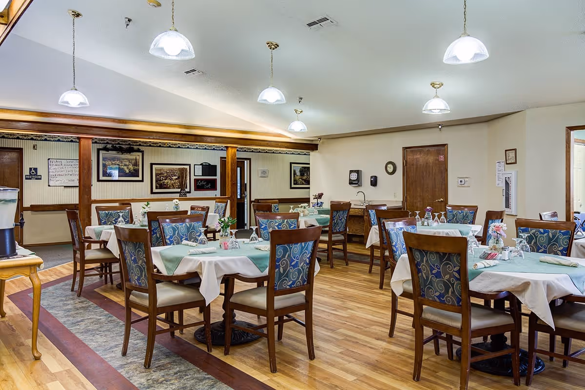 A dining room in a senior living facility with multiple tables covered with white and green tablecloths, each set with glasses, napkins, and small flower arrangements. The room has wooden floors, several hanging pendant lights, and framed artwork on the walls. There is a water dispenser on a small table to the left and a door labeled restroom in the background.