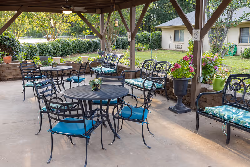 Covered outdoor patio area with several black metal tables and chairs with blue cushions, surrounded by potted plants and greenery, with a lawn and building visible in the background.