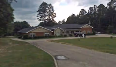 Single-story senior living facility with brick exterior, driveway, grassy lawn, and trees in background.