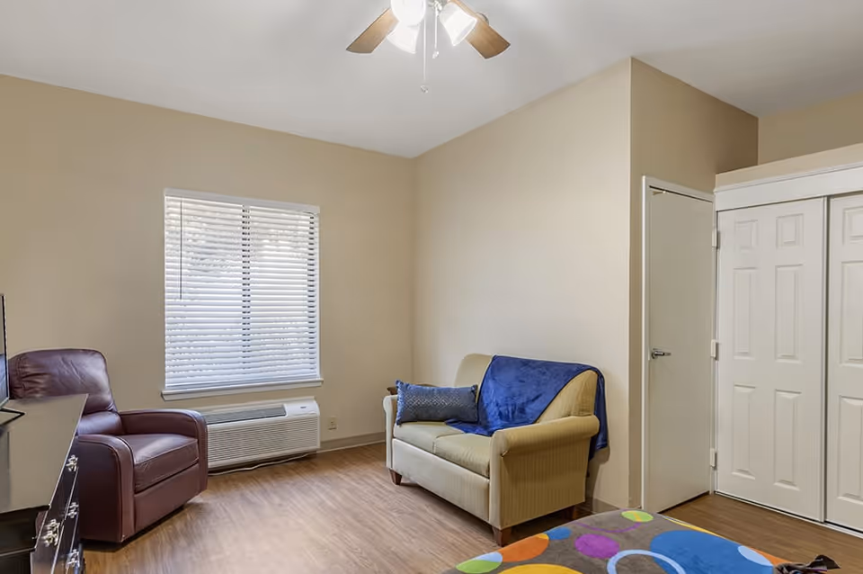 A cozy living room area with a beige loveseat covered partially with a blue throw blanket and a matching pillow, a brown leather armchair, a window with white blinds, a ceiling fan with lights, and a colorful rug on a wooden floor. There is a white closet door and a small door on the right wall.