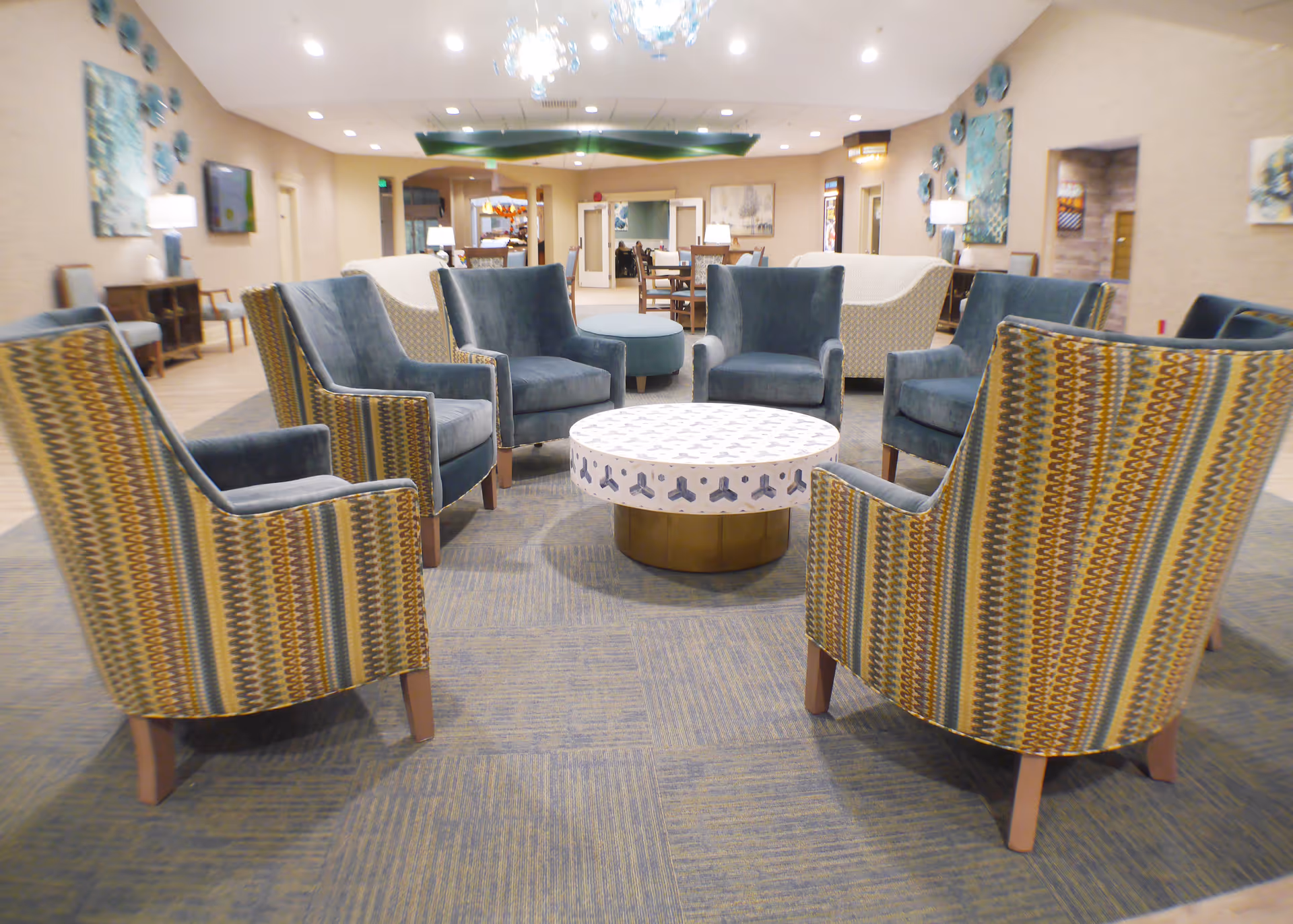 A spacious and well-lit lounge area with a circular arrangement of eight armchairs around a round patterned coffee table. The chairs have a mix of solid blue and striped upholstery. The room features a carpeted floor, modern lighting fixtures including a chandelier, and various pieces of wall art. In the background, there are additional seating areas and tables, suggesting a communal space within a senior living facility.