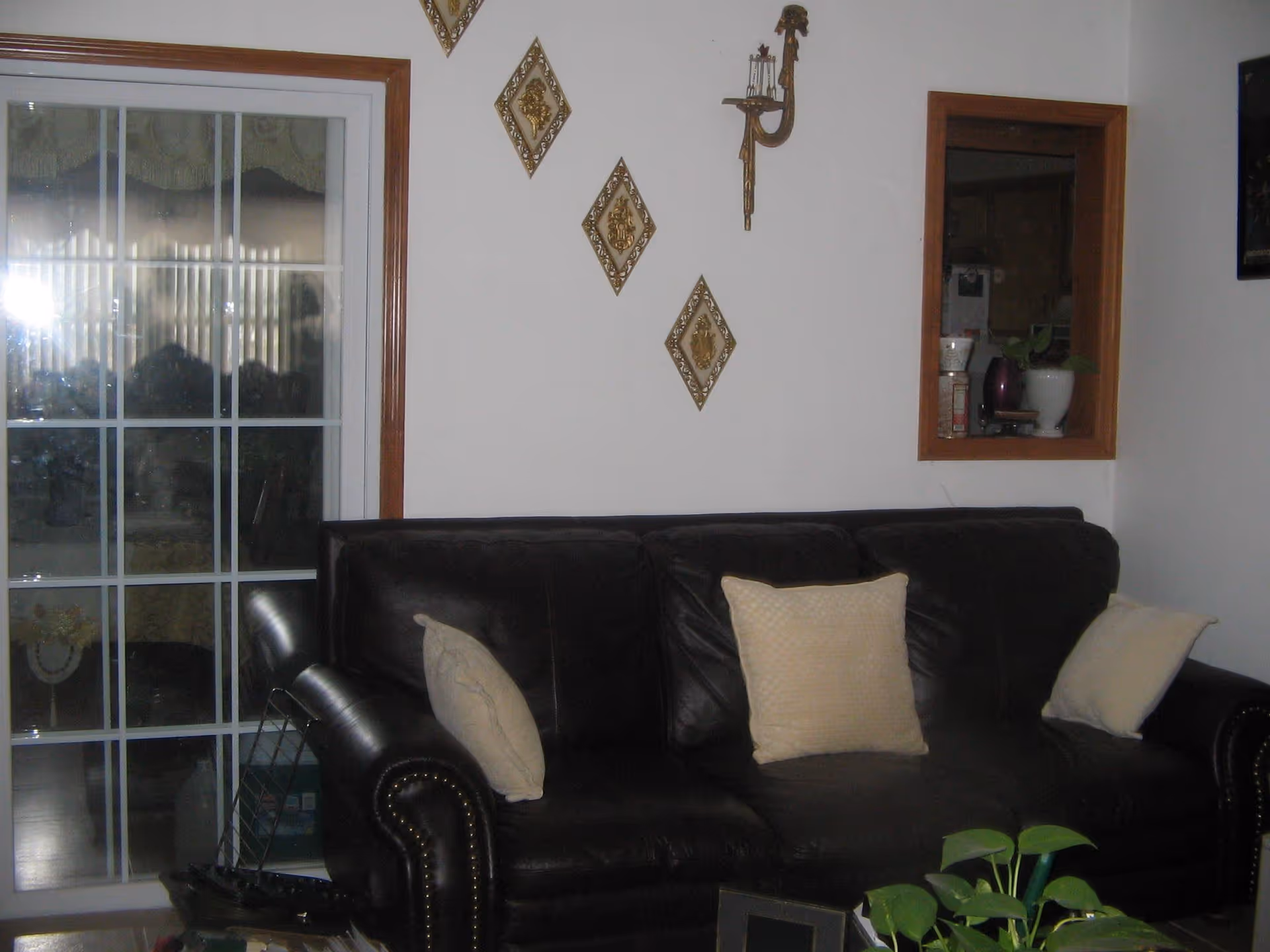 A living room area with a dark brown leather couch adorned with three beige pillows. Behind the couch is a white wall decorated with three diamond-shaped wall ornaments and a candle holder. To the left, there is a sliding glass door with a wooden frame, and to the right, a small open window-like shelf with a white pot and some items inside. A green plant is partially visible in the foreground.