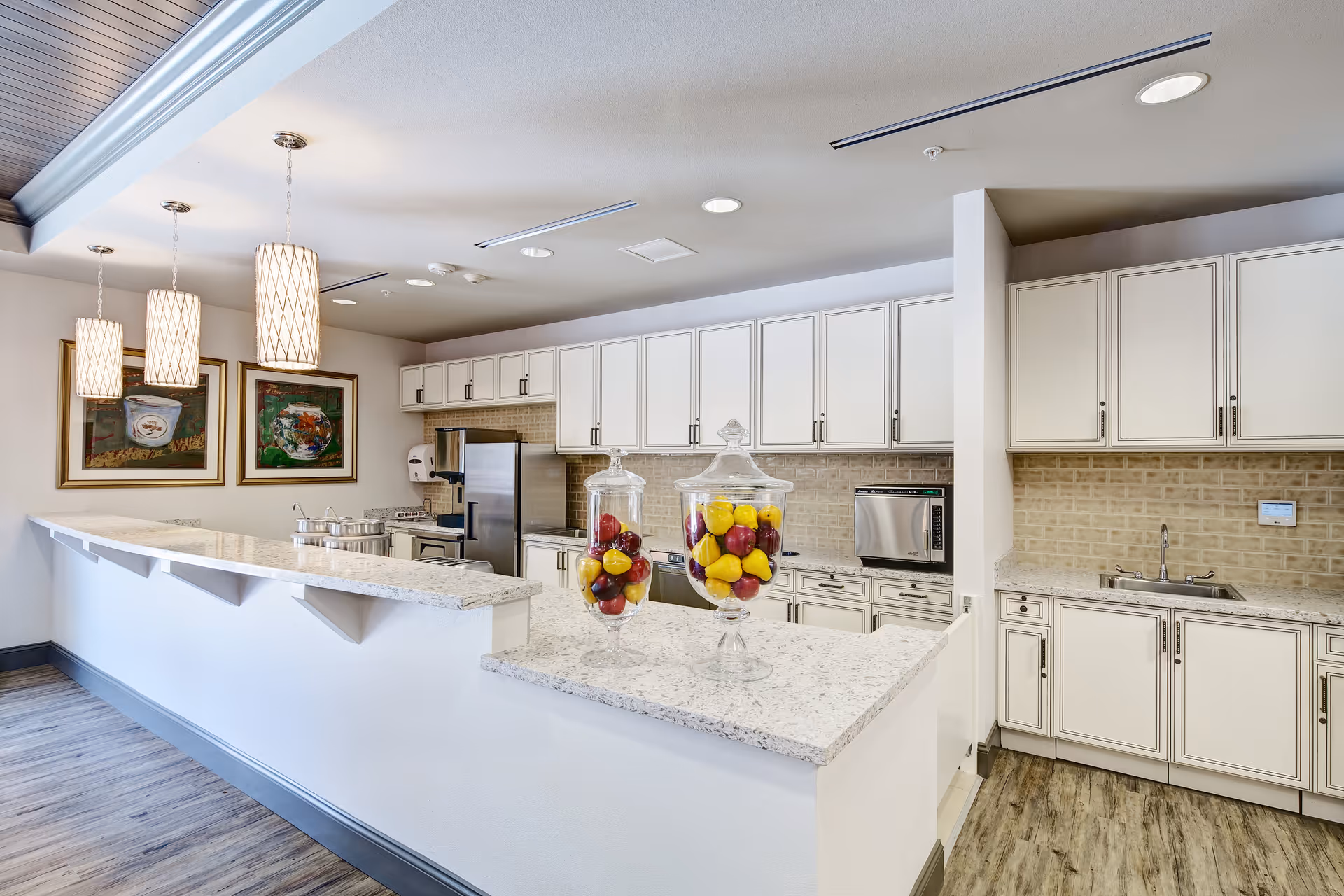 Bright communal kitchen with a long marble-topped serving counter, white cabinets, stainless appliances, pendant lights, and glass jars of fruit.