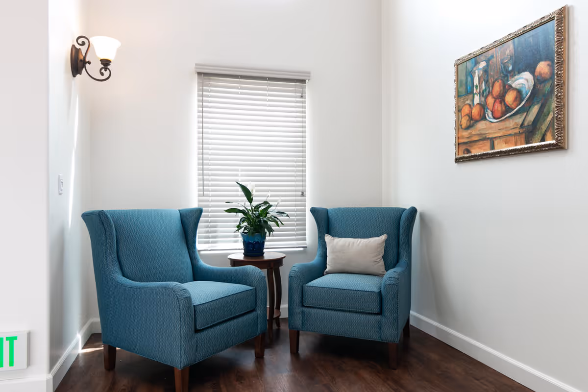 A cozy corner with two blue upholstered armchairs separated by a small round wooden table holding a potted plant. The wall behind has a window with closed blinds and a framed painting of fruit on the right side. A wall-mounted light fixture is on the left wall, and the floor is dark wood.