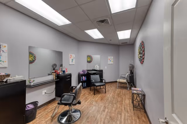 Interior view of a small salon or grooming room with two black salon chairs in front of large mirrors mounted on the wall. The room has light purple walls, wood flooring, and fluorescent ceiling lights. There are small cabinets and shelves with hair care products, a water cooler, and some decorative wall art.