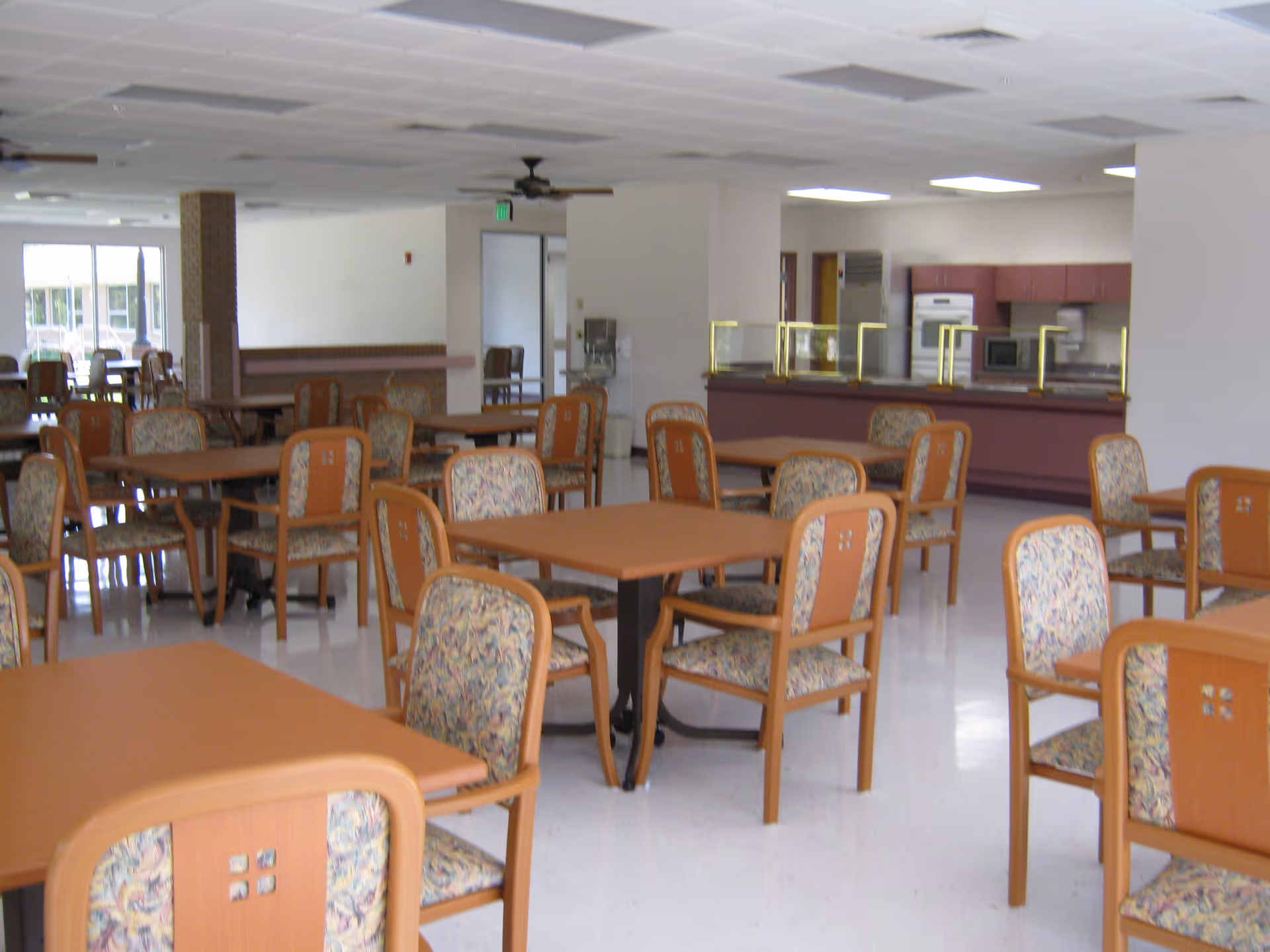 A spacious dining area with multiple wooden tables and chairs featuring floral-patterned cushions. The room has a white tiled floor, white walls, and a ceiling with recessed lighting and ceiling fans. In the background, there is a kitchen area with appliances and a serving counter with glass partitions.