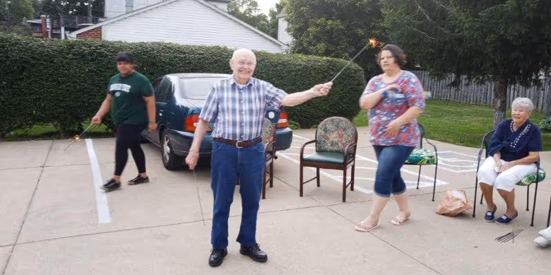 Four people outdoors on a paved area near parked cars and greenery. An elderly man in a plaid shirt and blue pants is holding a lit sparkler and smiling. A woman in a floral top and capri pants is walking nearby, and another person in a green shirt is holding a sparkler in the background. An elderly woman is seated on a chair with a bag on the ground beside her, smiling and watching the others.