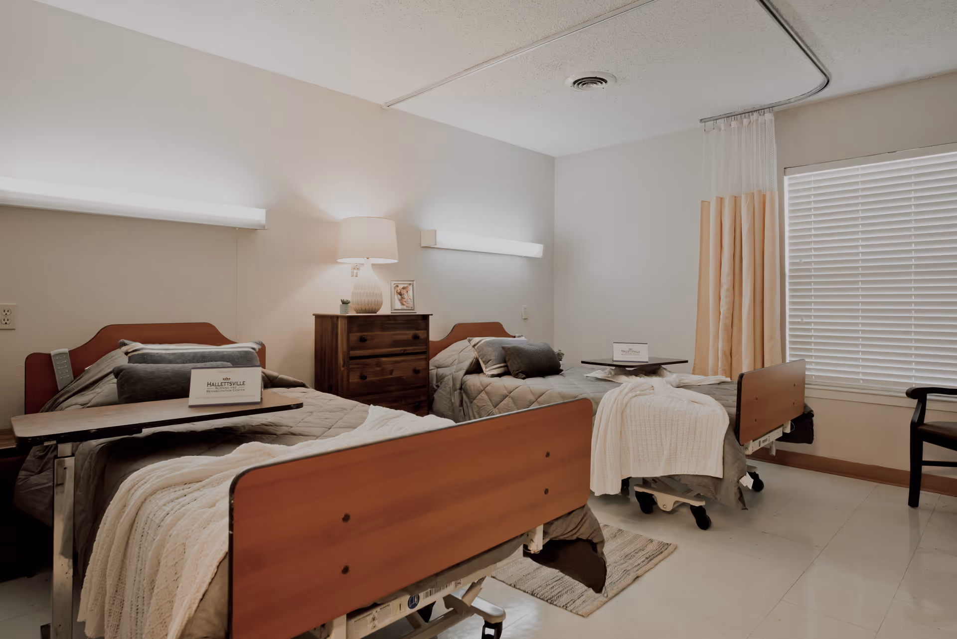 A nursing facility room with two hospital beds, each made with beige and gray bedding and a pillow. Between the beds is a wooden dresser with a lamp and a framed photo on top. Each bed has an overbed table with a sign that reads Hallettsville Nursing and Rehabilitation Center. The room has a window with closed blinds and a privacy curtain on a ceiling track. There is a wooden chair near the window.