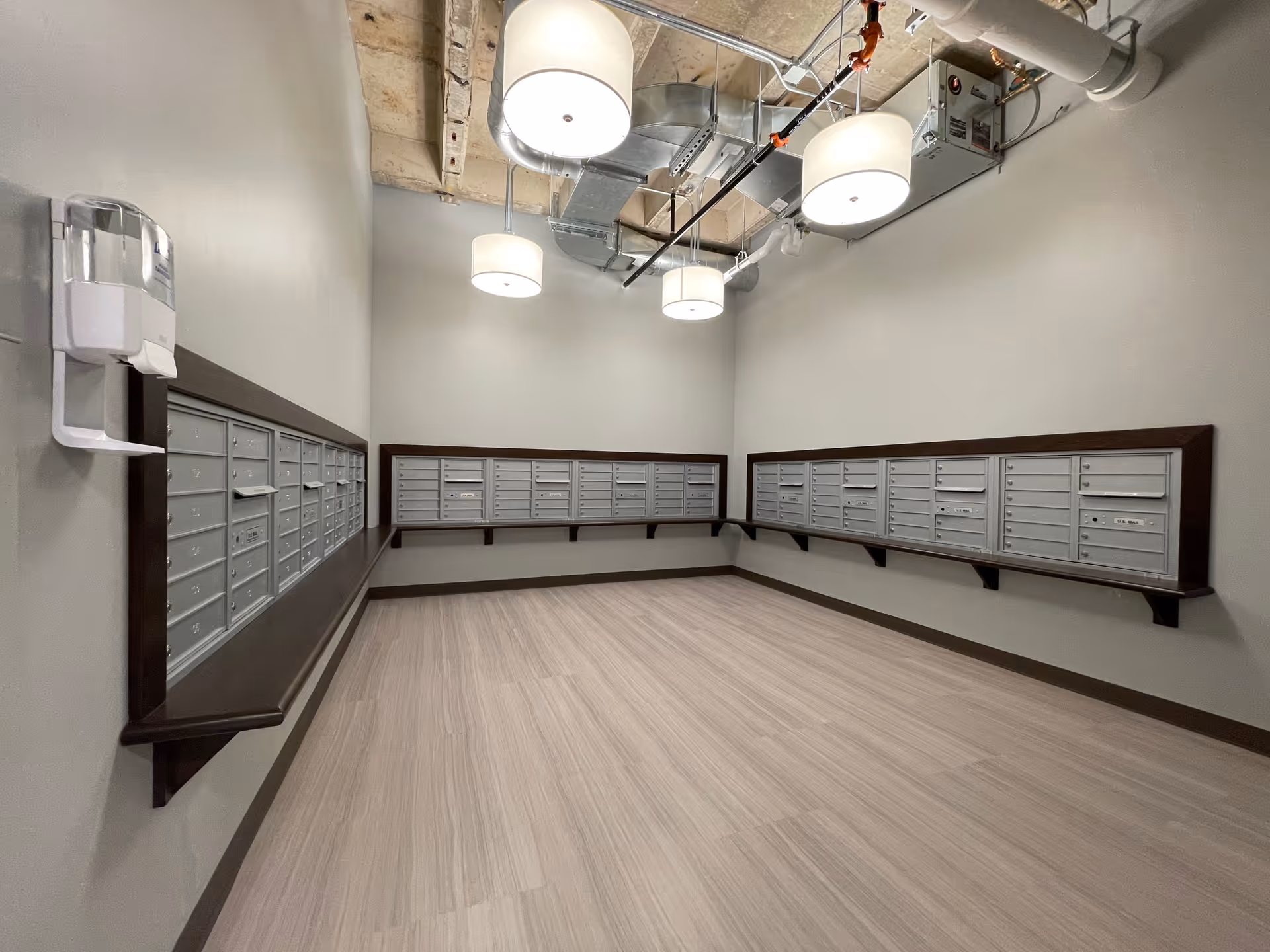 A room with multiple rows of silver mailboxes mounted on two adjacent walls, with a light wood floor and industrial-style ceiling with exposed pipes and four round ceiling lights.