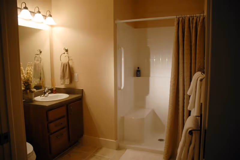 A bathroom with a wooden vanity featuring a sink and mirror above it, a towel hanging on a ring, and a shower area with a built-in seat and a beige shower curtain. The bathroom is softly lit with wall-mounted lights above the mirror.