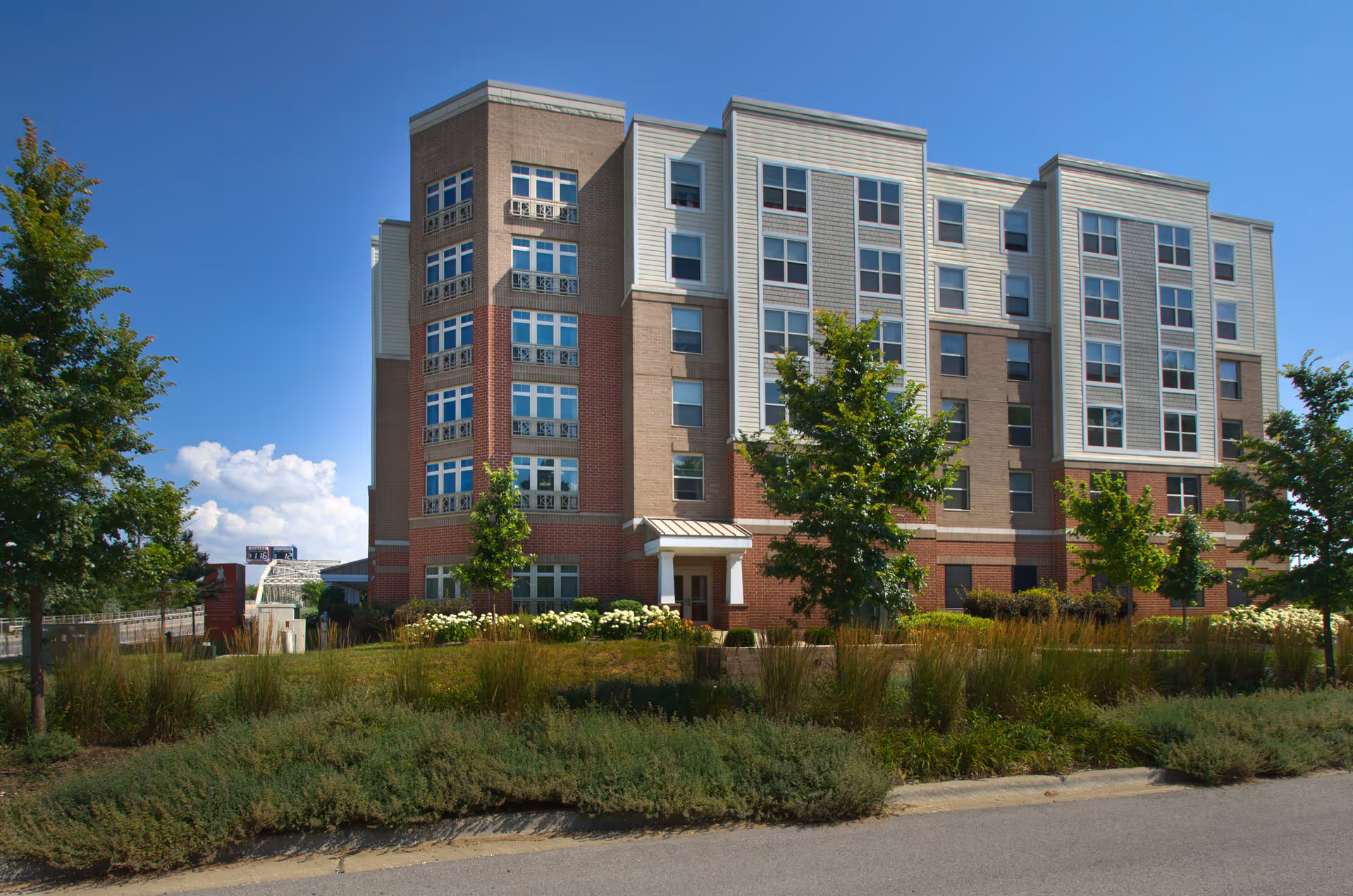 Exterior view of a multi-story senior living facility building with a mix of brick and siding facade, surrounded by landscaped greenery and trees under a clear blue sky.