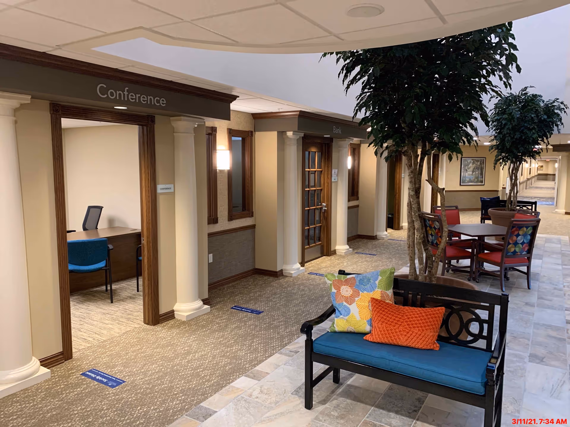 Interior hallway of Creek Valley Retirement Community featuring a seating area with a bench adorned with colorful pillows, potted trees, and tables with chairs. Doors labeled Conference and Bank are visible along the hallway.