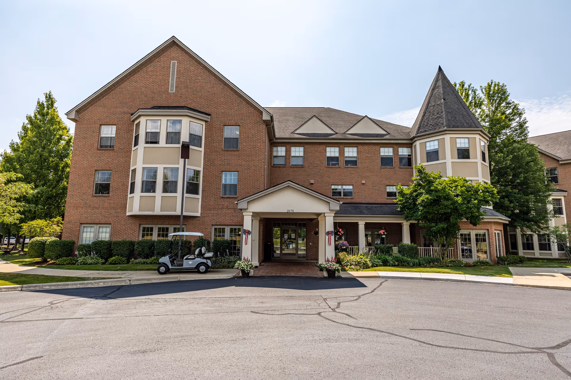 Front view of Lourdes Senior Community featuring a brick facade, large windows, and a welcoming entrance with potted plants and a golf cart parked nearby.