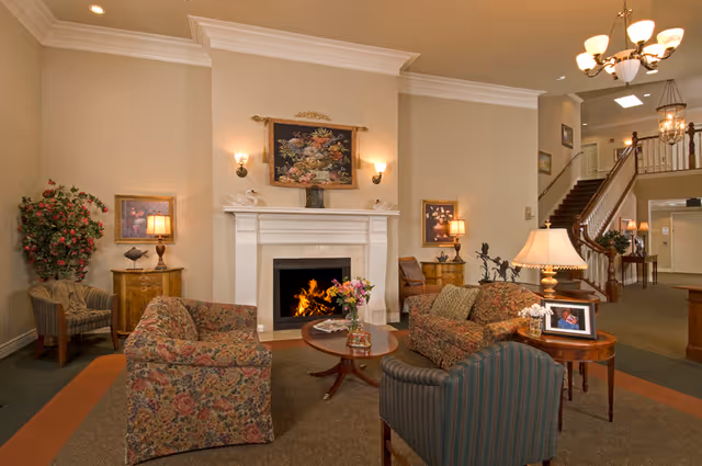 A cozy living room area with floral patterned sofas and a striped armchair arranged around a wooden coffee table with a vase of flowers. There is a white fireplace with a fire burning, flanked by two wall sconces and decorative artwork above. The room has warm lighting, a large plant in the corner, and a staircase leading to an upper floor in the background.