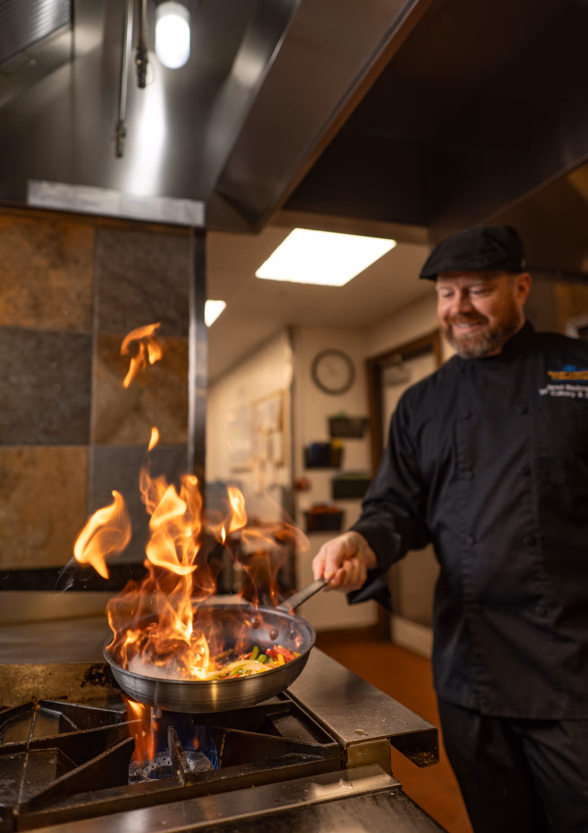 A chef wearing a black uniform and hat is cooking with a frying pan over a gas stove, with flames rising from the pan in a commercial kitchen setting.