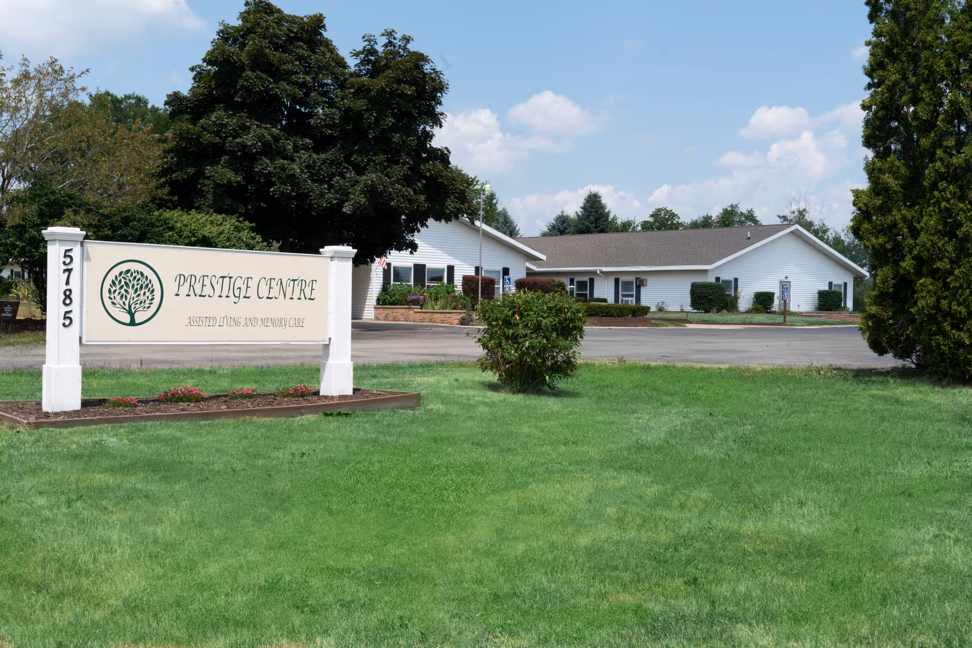 Exterior view of a single-story white building with a gray roof, surrounded by green grass, trees, and bushes under a partly cloudy sky. In the foreground, there is a large sign that reads 'Prestige Centre Assisted Living and Memory Care' with the number 5785 on the left post.