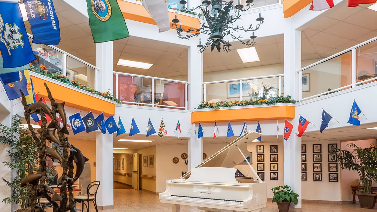Interior view of a senior living facility with a white grand piano in the center of a spacious, two-story atrium. The area is decorated with multiple state flags hanging from the upper level railing, green plants, and a large chandelier overhead. The walls feature framed pictures and the space has a bright, welcoming atmosphere.