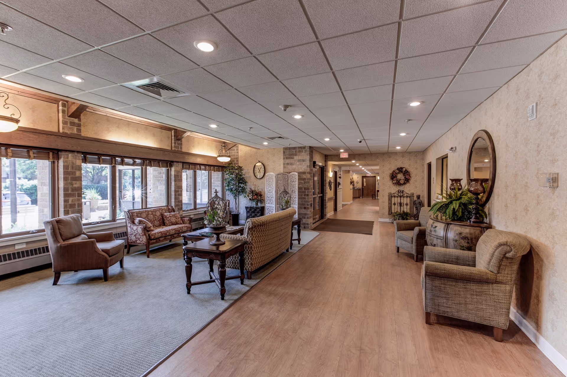 A spacious and well-lit senior living facility common area with comfortable seating including armchairs and sofas arranged around a coffee table. Large windows on the left side allow natural light to fill the room. The walls are decorated with plants, a clock, and a decorative screen. The floor transitions from carpet in the seating area to wood laminate in the hallway leading further into the facility.
