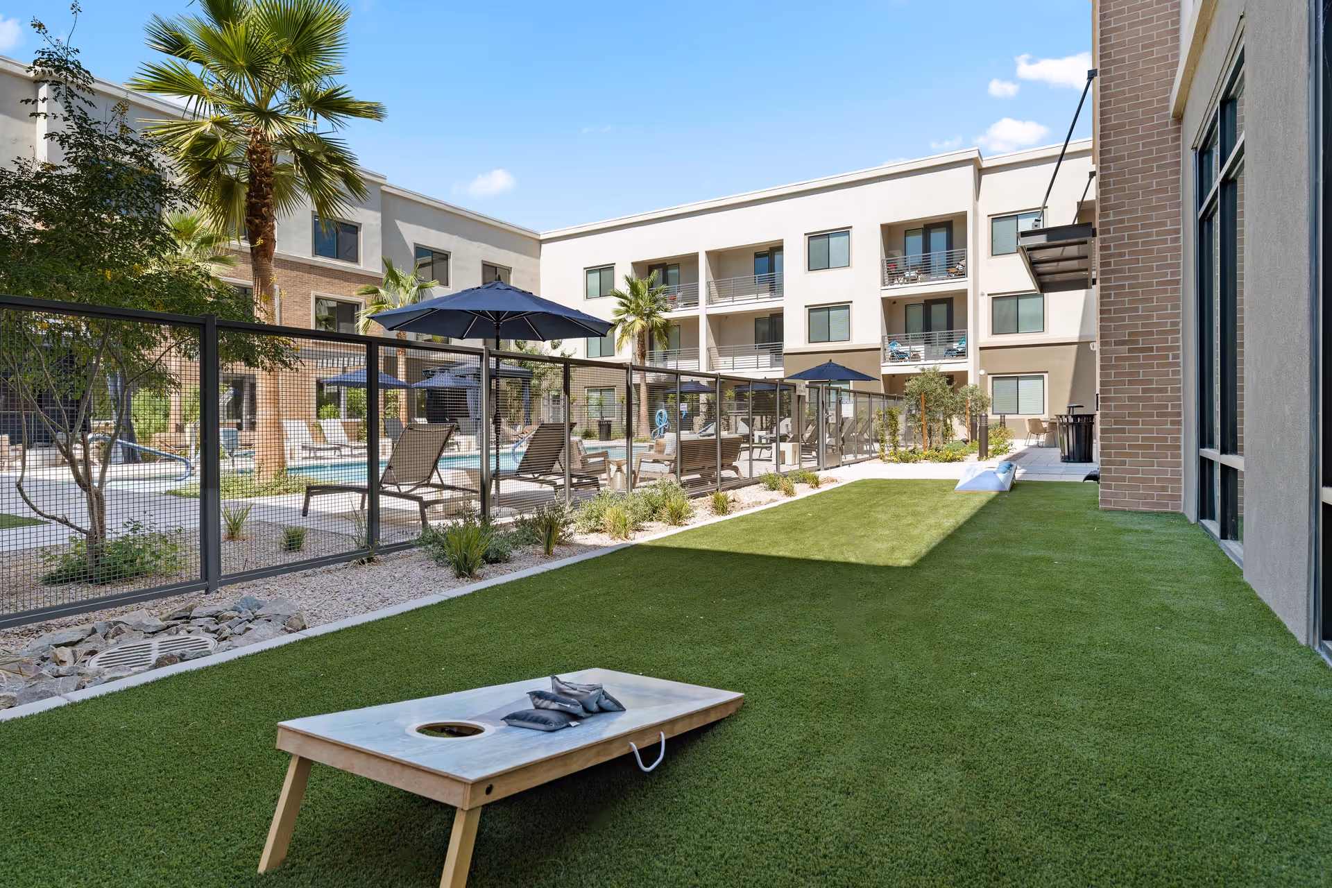 Outdoor courtyard area of a senior living facility with artificial grass, a cornhole game set up, lounge chairs, umbrellas, palm trees, and a fenced swimming pool surrounded by a three-story building under a clear blue sky.