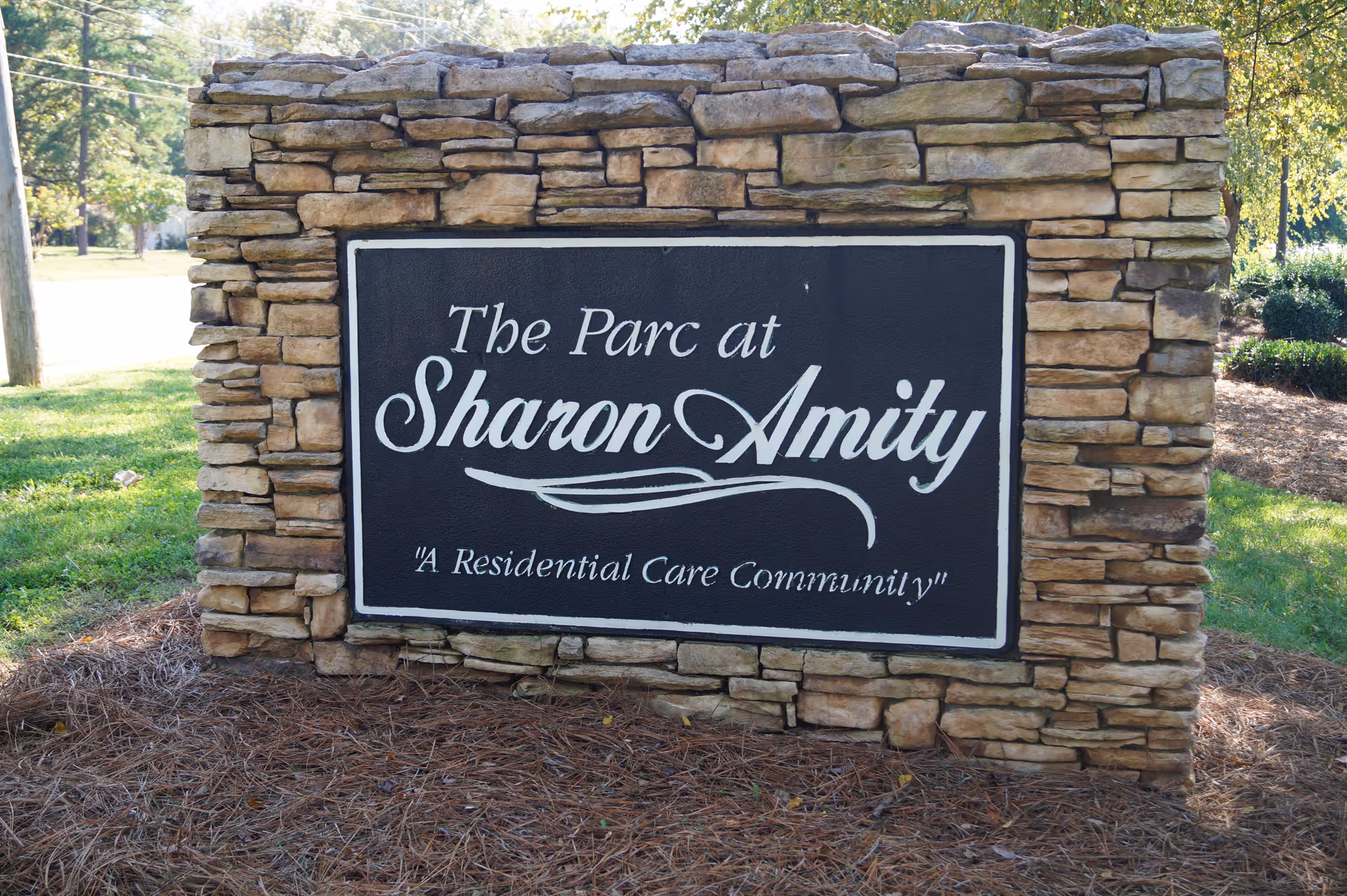 Stone monument sign with a black plaque that reads 'The Parc at Sharon Amity, A Residential Care Community' surrounded by grass, pine straw, and trees in the background.