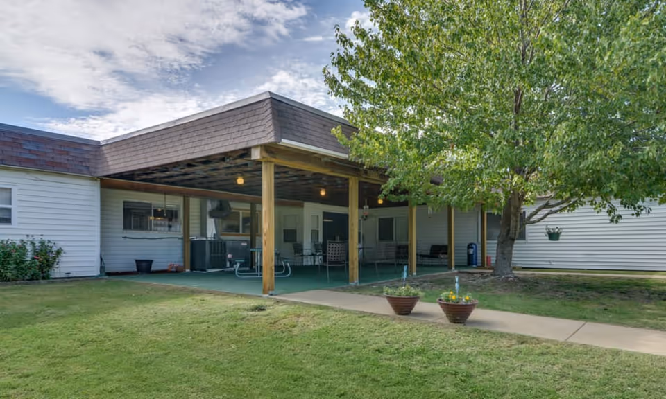Outdoor covered patio area with seating and a picnic table at a nursing center. The patio is supported by wooden posts and has a green floor covering. There is a tree with green leaves nearby and two flower pots on a concrete walkway leading to the patio. The building exterior is white with windows and a brown shingled roof.
