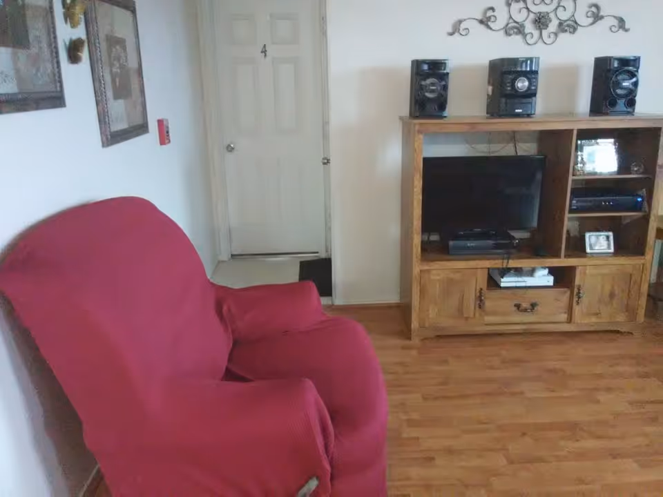 A living room with a red armchair covered by a slipcover on the left side, a wooden entertainment center with a TV, stereo system, and framed photos on the right side, and a white door with the number 4 in the background.