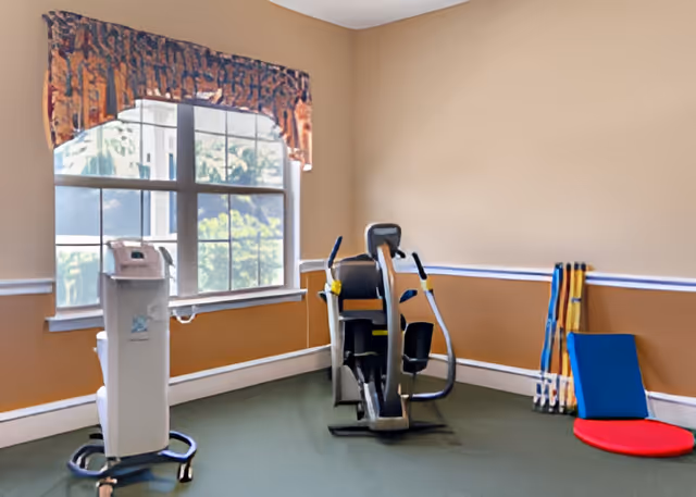 A small exercise room with a large window covered by a patterned valance. The room contains a piece of exercise equipment, a medical device on wheels, and some exercise mats and poles stacked against the wall.