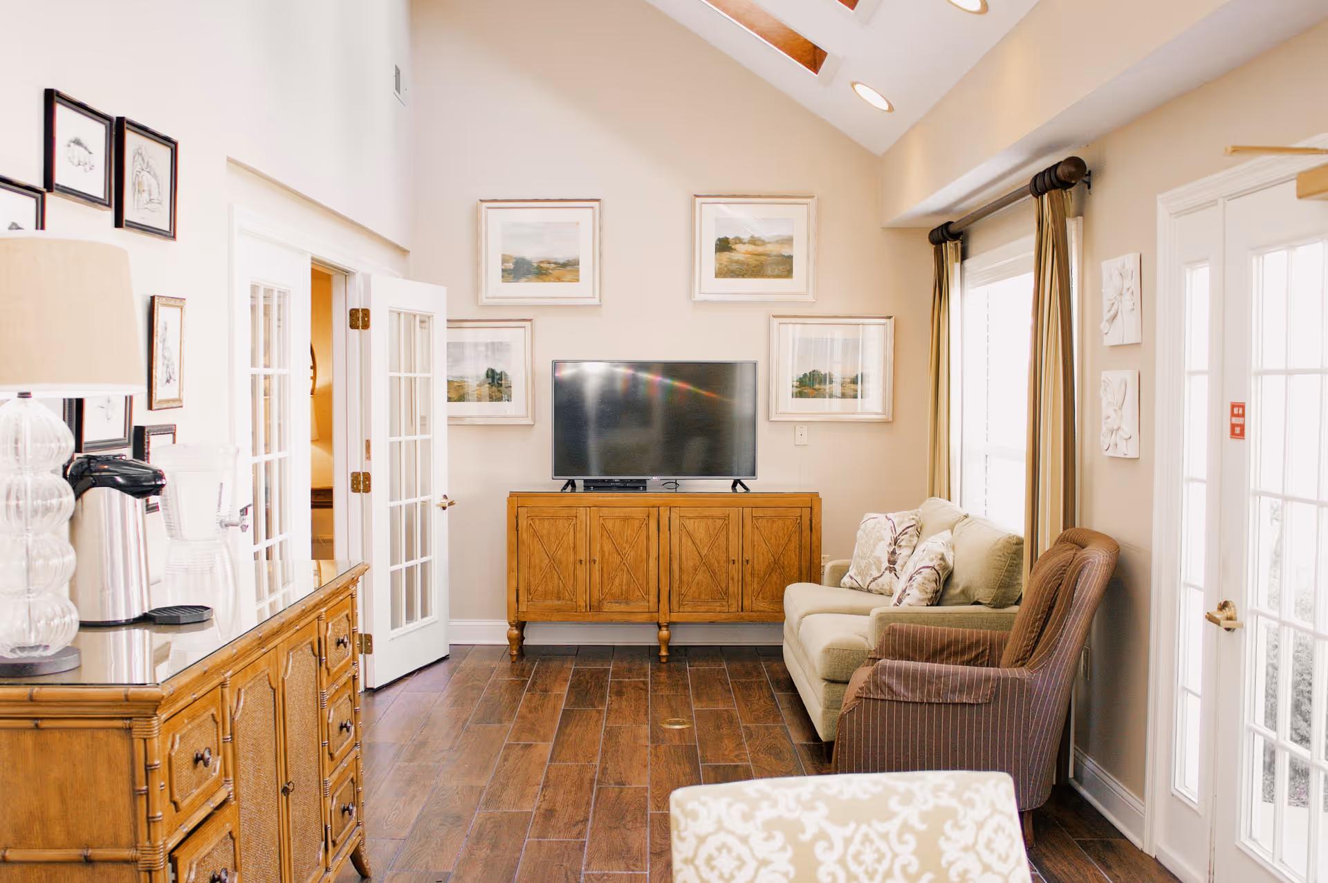 A bright living room with wooden flooring, a beige sofa with patterned cushions, a brown armchair, a wooden TV stand with a flat-screen TV, and framed landscape paintings on the wall. There is a wooden sideboard with a lamp, coffee maker, and glass pitcher. French doors and large windows with curtains allow natural light to fill the room.