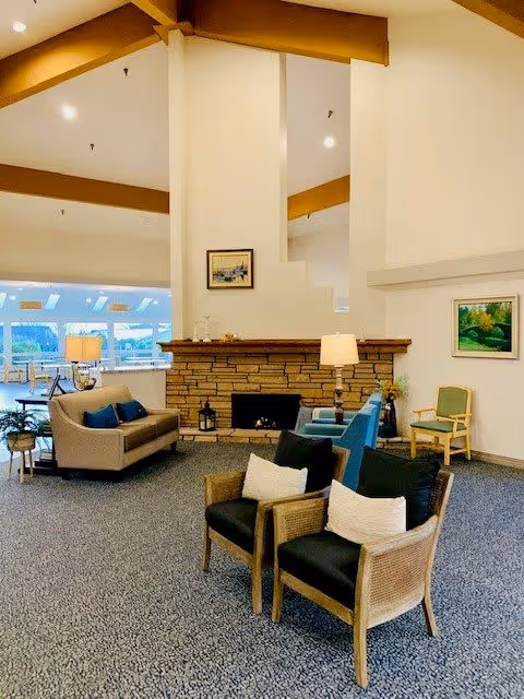 A spacious living room area in a retirement community featuring a stone fireplace in the center with a painting above it. The room has multiple seating options including two wicker chairs with cushions in the foreground, a beige sofa with blue pillows to the left, and a green armchair with a wooden frame to the right. There are two table lamps providing warm lighting, and large windows in the background letting in natural light.