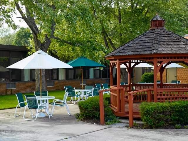 Outdoor seating area with white and green umbrellas shading tables and chairs, surrounded by green trees and bushes. A wooden gazebo with a shingled roof is visible on the right side. The background shows a brick building with windows.