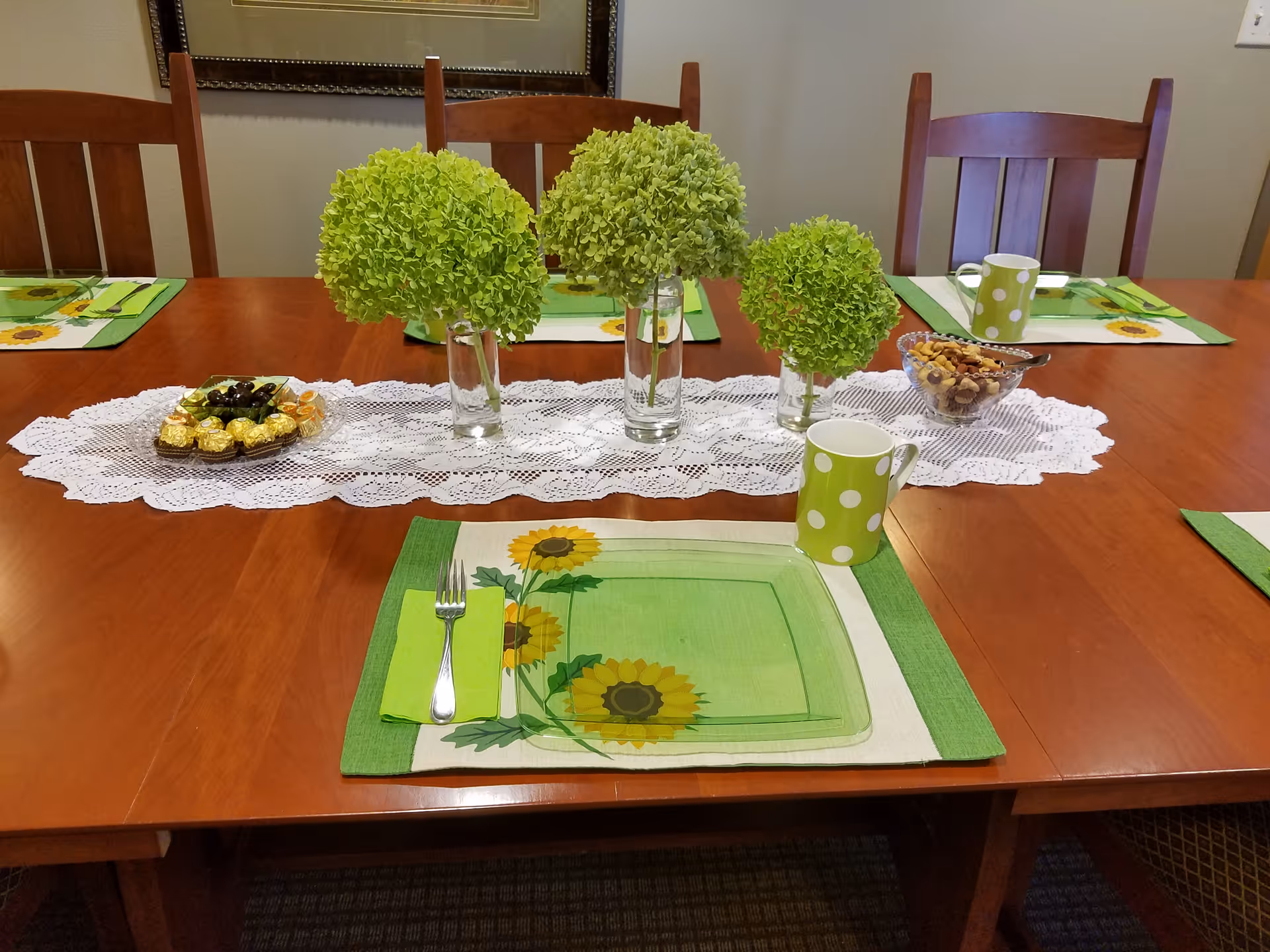 A wooden dining table set with green sunflower-themed placemats, green polka dot mugs, and silverware wrapped in green napkins. The table has a white lace runner with three glass vases holding green hydrangea flowers. There is a bowl of mixed nuts and a plate with chocolates and olives on the table. Four wooden chairs are visible around the table.