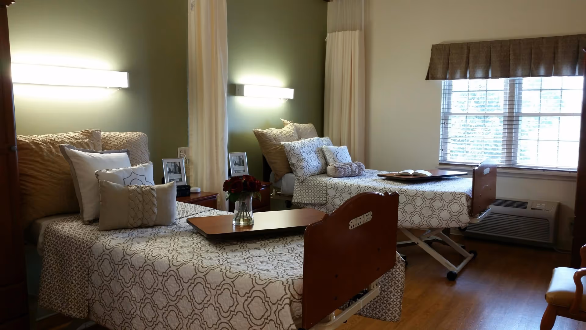 A well-lit room with two hospital-style beds, each made up with patterned bedspreads and multiple pillows. There is a wooden tray on each bed, one holding a vase with red flowers and the other an open book. The room has a large window with blinds and a valance, wooden flooring, and a green accent wall with a wall-mounted light above each bed. A curtain divider is partially visible between the beds.