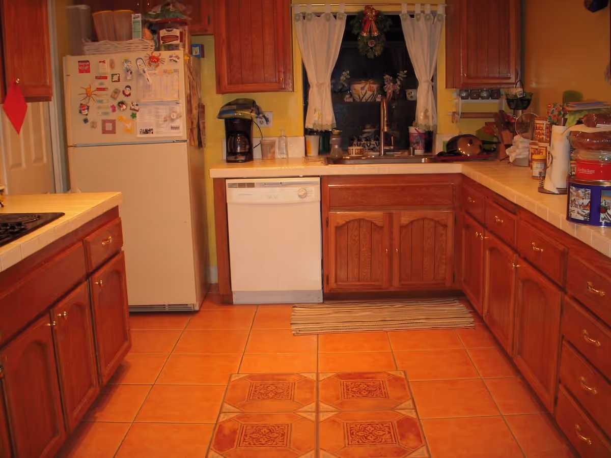 A kitchen with wooden cabinets, a white refrigerator covered with magnets and papers, a dishwasher, a coffee maker, a sink under a window with white curtains, and various kitchen items on the countertops. The floor is tiled with orange and patterned tiles.