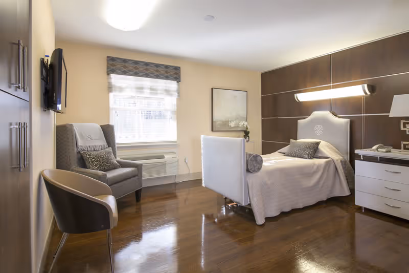 A nicely furnished senior living room featuring a bed, chair, and dresser with modern decor.