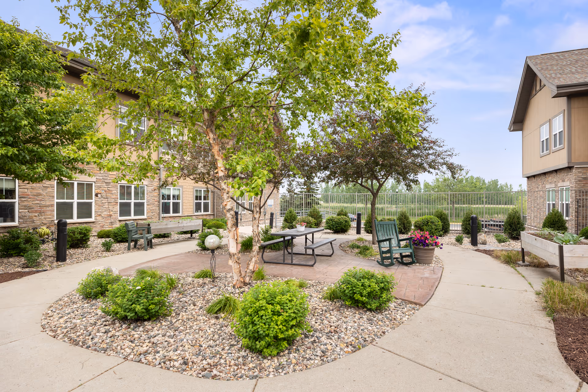 Sunny courtyard patio with trees, shrubs, a picnic table, benches and rocking chairs between residential buildings.
