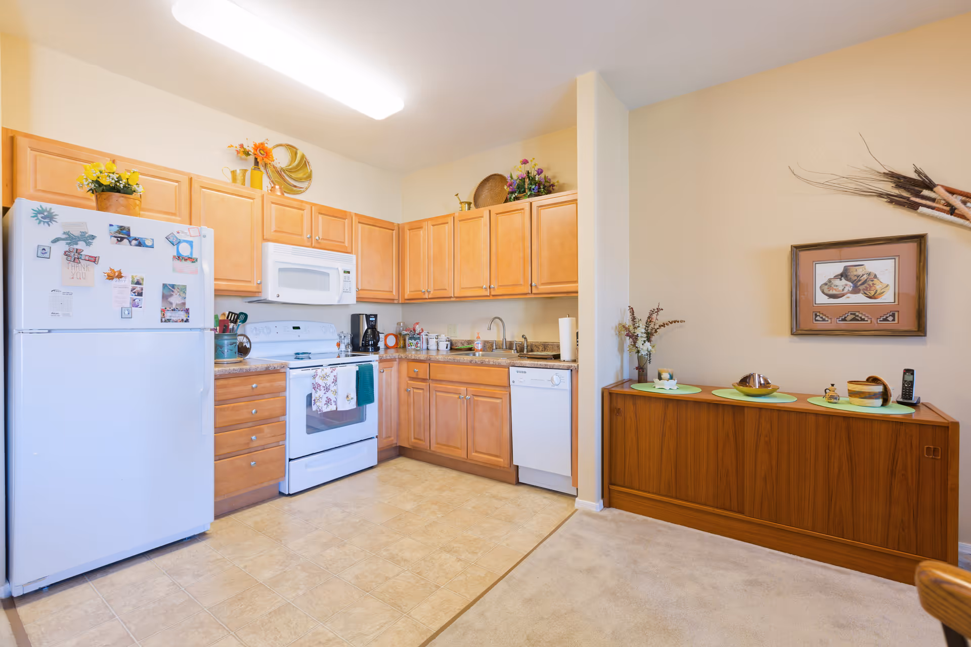 A bright kitchen area with light wood cabinets, a white refrigerator covered with magnets and photos, a white stove with a microwave above it, a coffee maker, a dishwasher, and a sink. The floor is tiled in the kitchen area and carpeted in the adjacent space. There is a wooden sideboard against the wall with decorative items and a framed picture hanging above it.