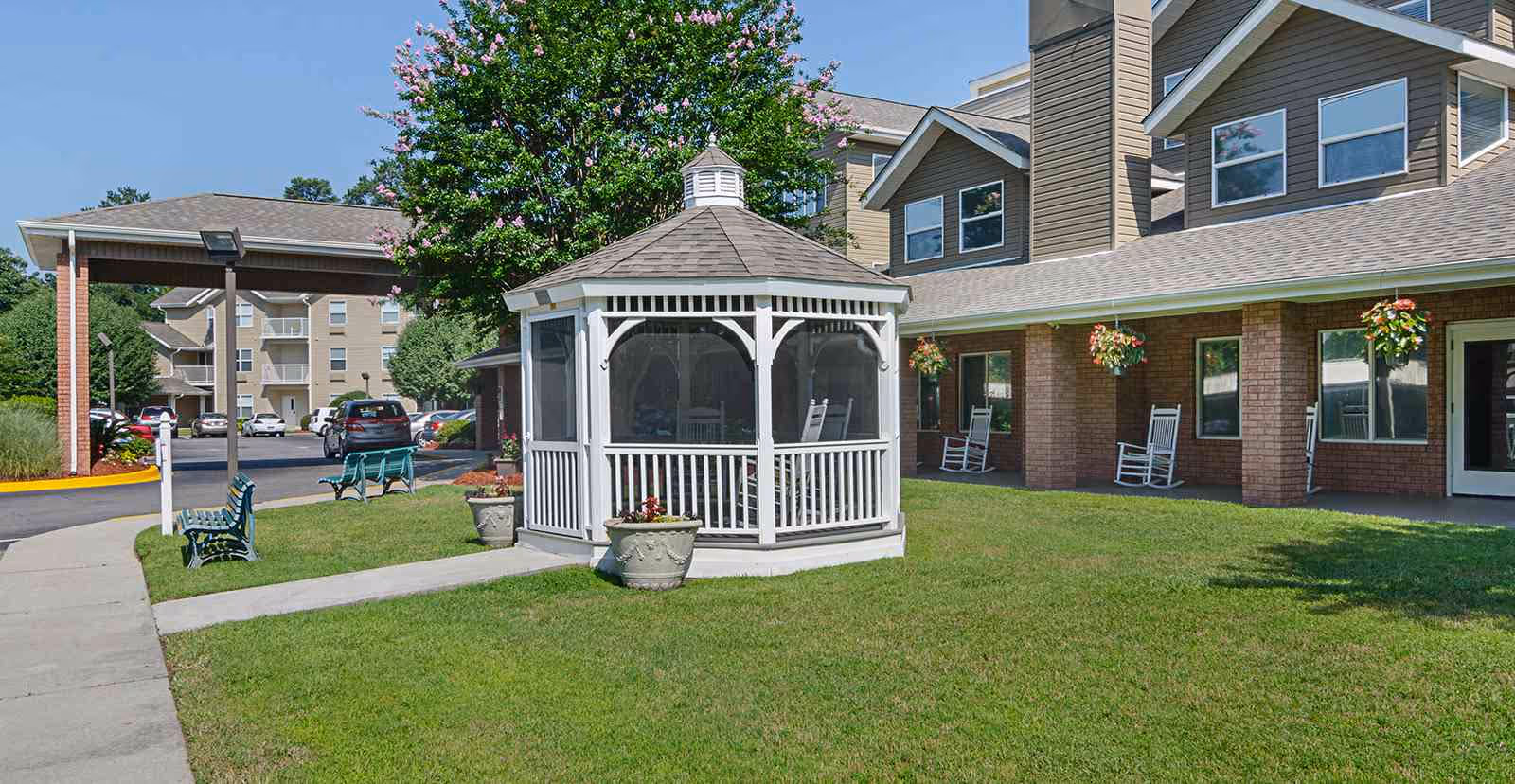 Outdoor area of Holiday Columbia Pines featuring a white gazebo with rocking chairs inside, surrounded by green grass and potted plants. The building exterior has brick walls, multiple windows, hanging flower baskets, and a covered porch with additional rocking chairs. There are benches along a sidewalk and parked cars in the background under a clear blue sky.