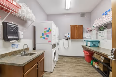 A clean laundry/utility room with stacked washers and dryers, a sink and cabinets, shelving with supplies, and a folding counter.